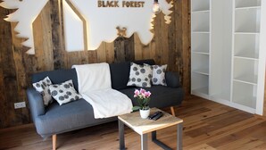 Een moderne woonkamer met een grijze bank, zwart-witte kussens, een witte plaid, een kleine tafel met een potplant en afstandsbediening, en een wit wandrek.