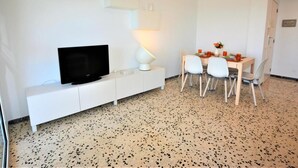 A modern room with a flat-screen TV on a white cabinet, a dining table with chairs, and a small white lamp. - Leonor 3º2ª - Calafell (Calafell)
