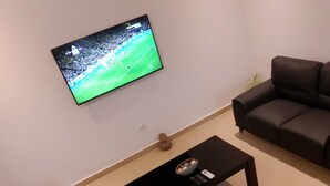 A living room with a flat-screen TV mounted on the wall displaying a soccer game, a black coffee table with a magazine and remote control, and a dark-colored sofa with a yellow pillow. - R&Rhome appartement meublé (Cotonou)