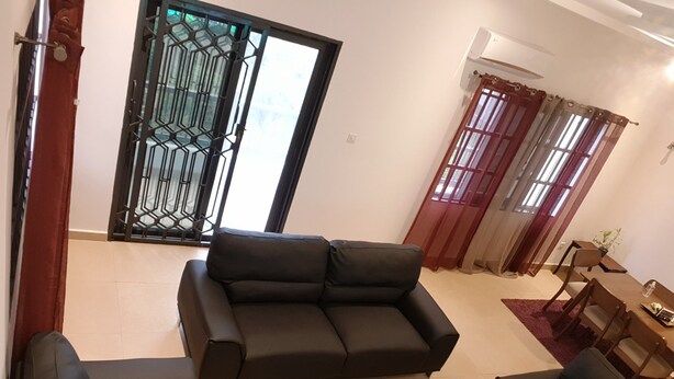 A living room with a black sofa, a coffee table, and a dining area with a table and chairs. - R&Rhome appartement meublé (Cotonou)