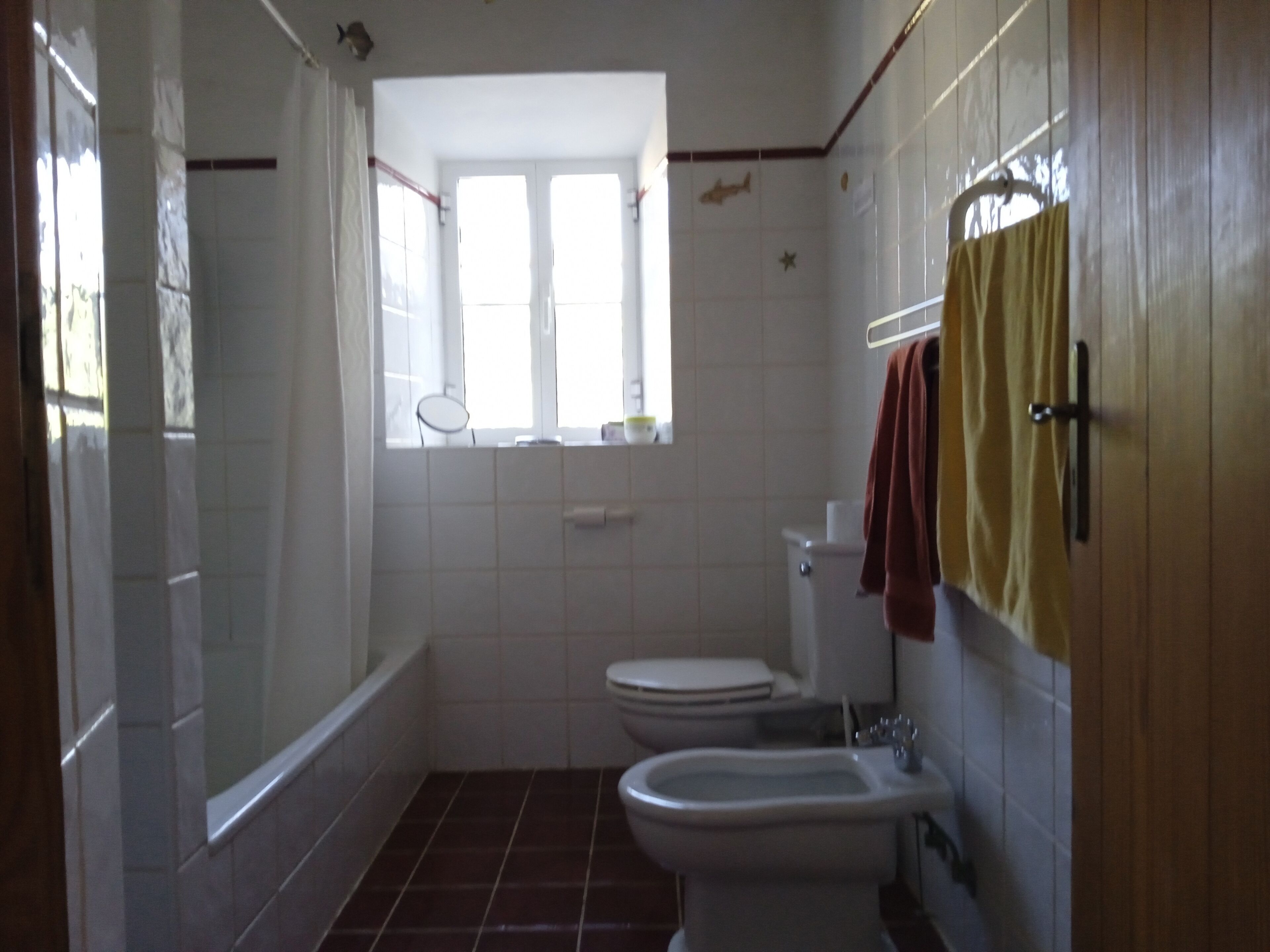 A small bathroom with a toilet, bidet, and bathtub. There is a window with curtains and a towel hanging on the wall.