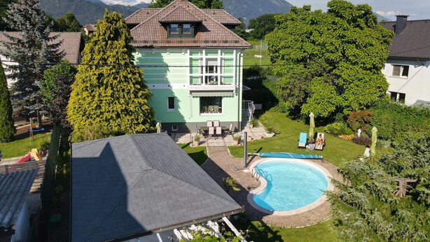 Ein zweistöckiges Haus mit Schwimmbad, umgeben von üppiger Vegetation und Bergen im Hintergrund.