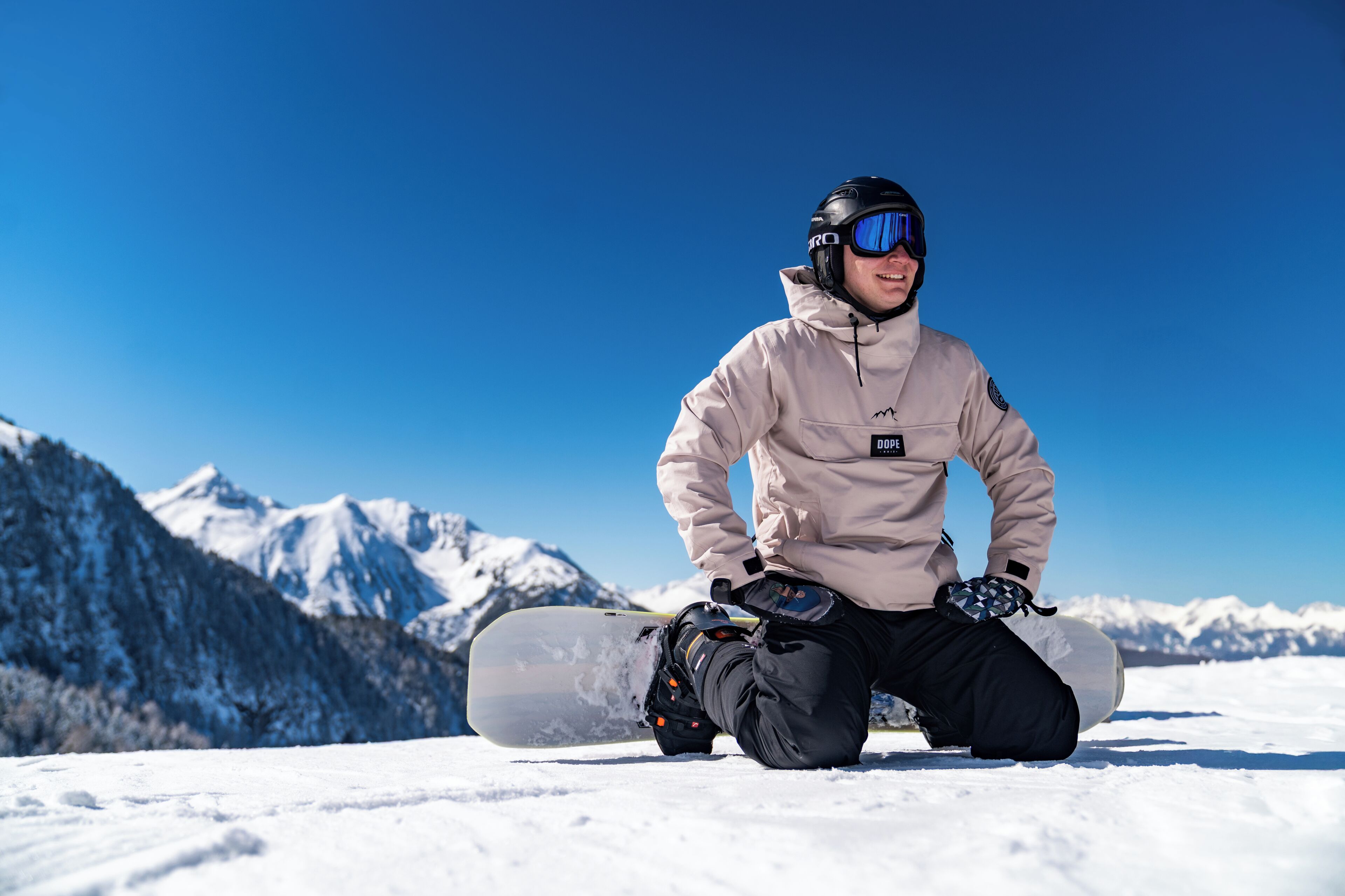 A person kneeling on snow with a snowboard, wearing a helmet and goggles, with snow-covered mountains in the background.