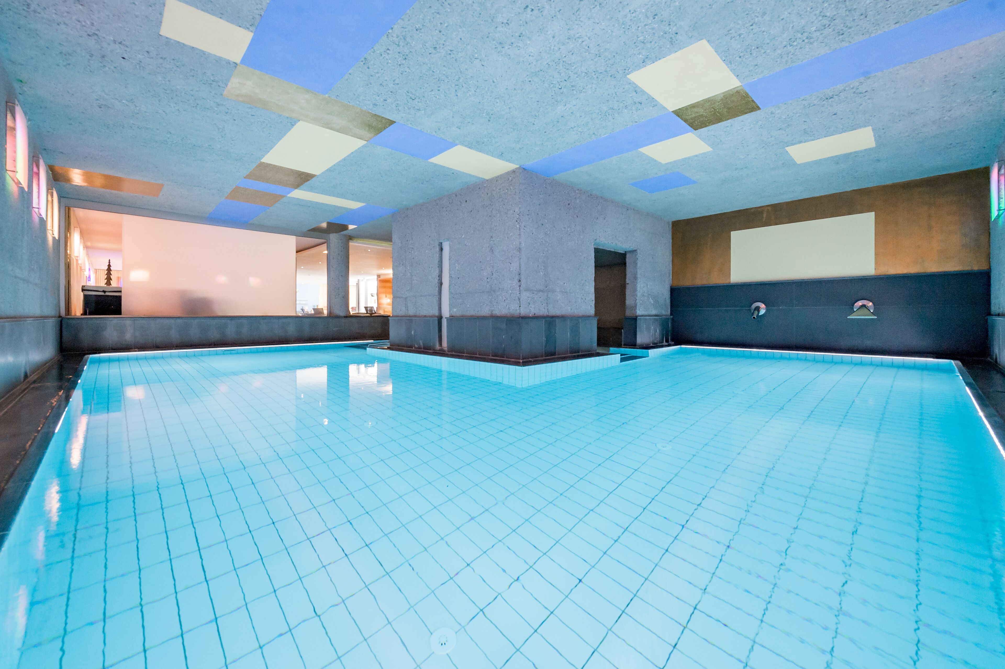 A large indoor swimming pool with a tiled floor and walls, a ceiling with geometric patterns, and a visible door leading to another room.