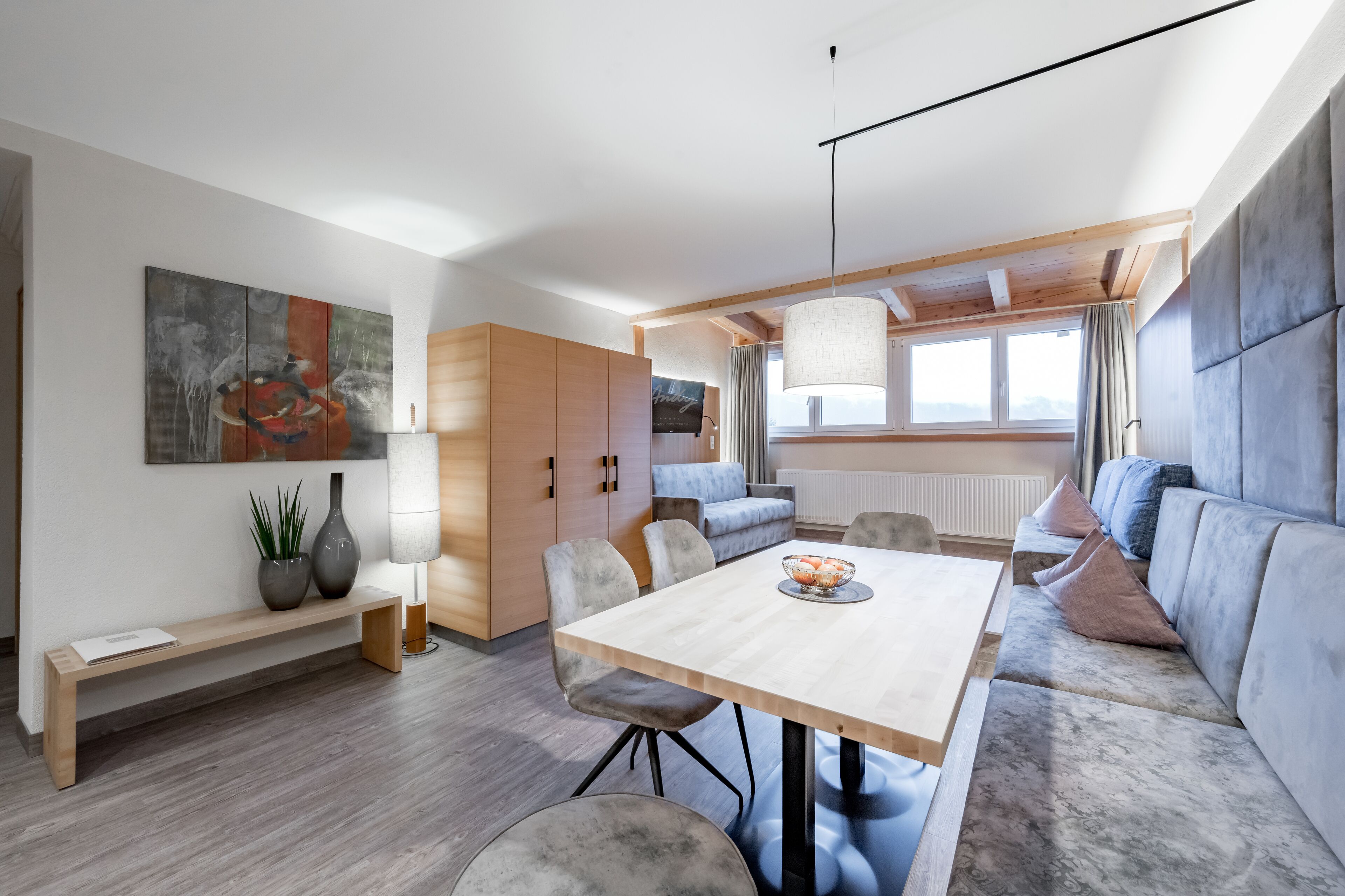 A modern living room with a wooden dining table, grey sofa, and a large window with curtains.
