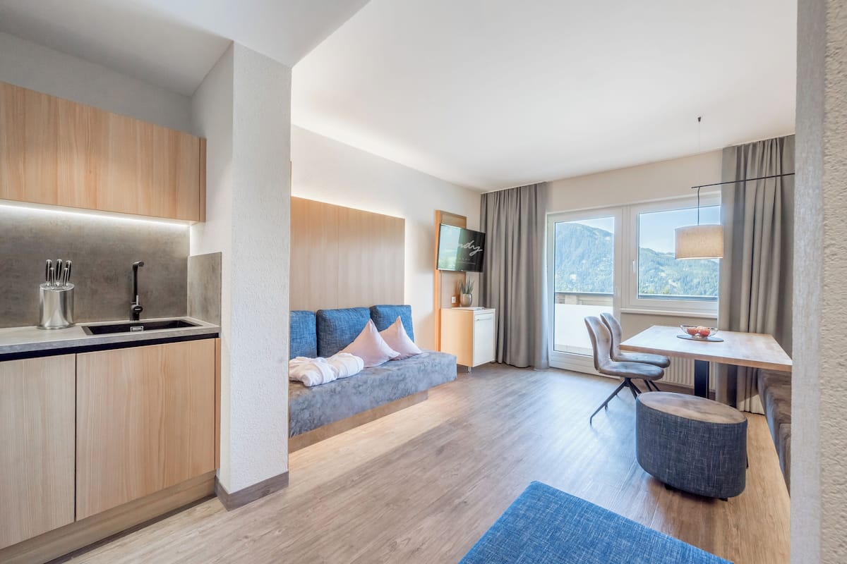 Quarto de hotel moderno com cozinha compacta, sofá, mesa de jantar e vista para as montanhas.
