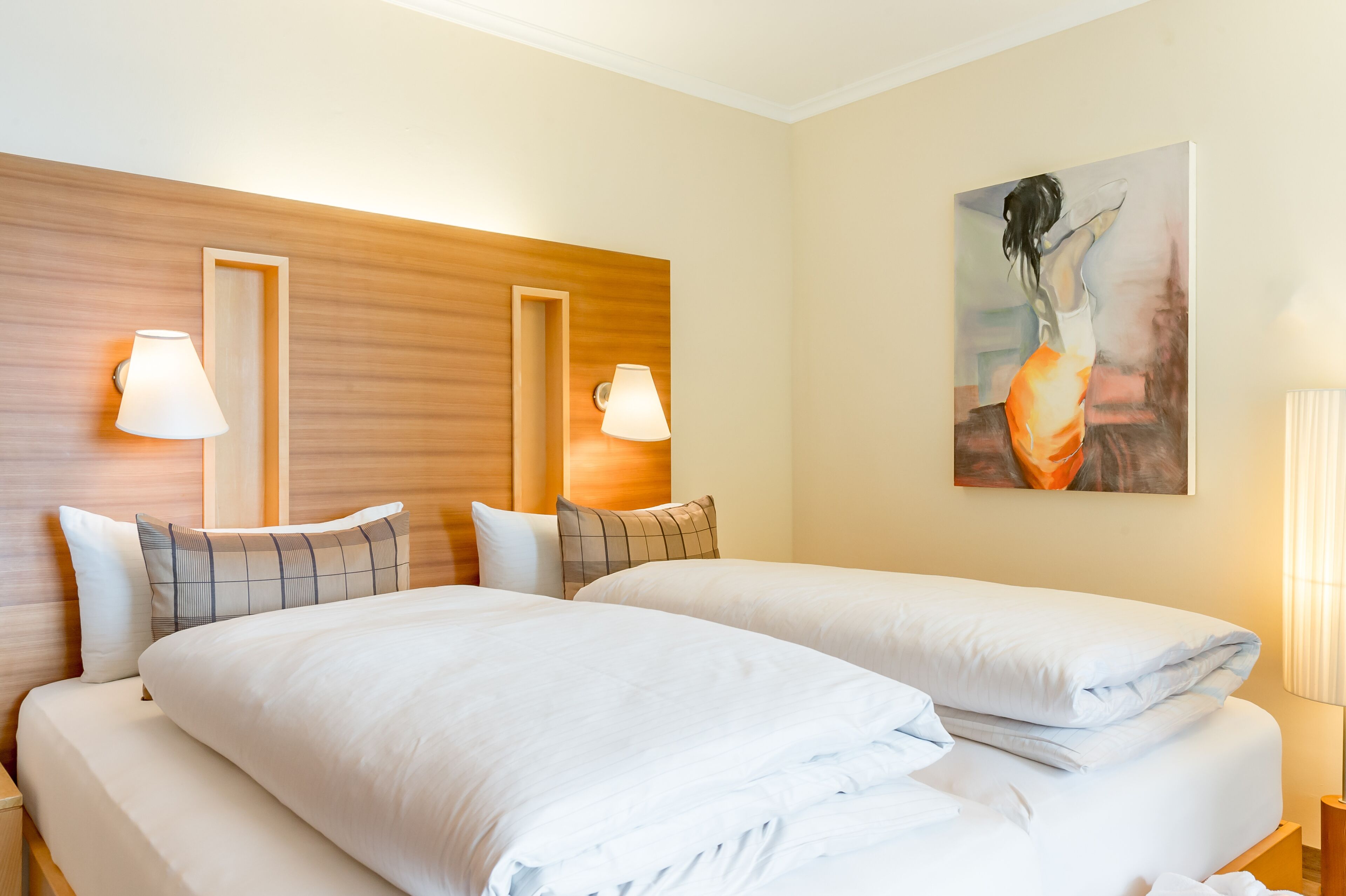 A hotel room with two beds, wooden headboard, wall-mounted lamps, and a painting.