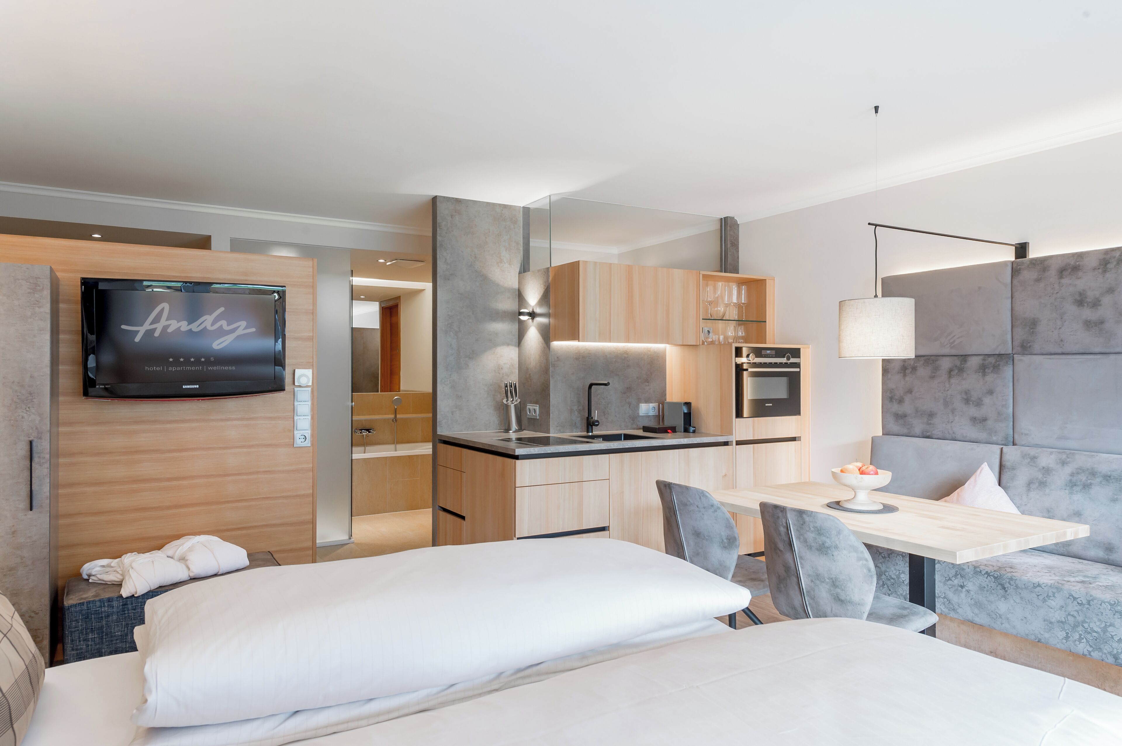 A modern hotel room with a bed, a dining table, a sofa, a TV, and a kitchenette.