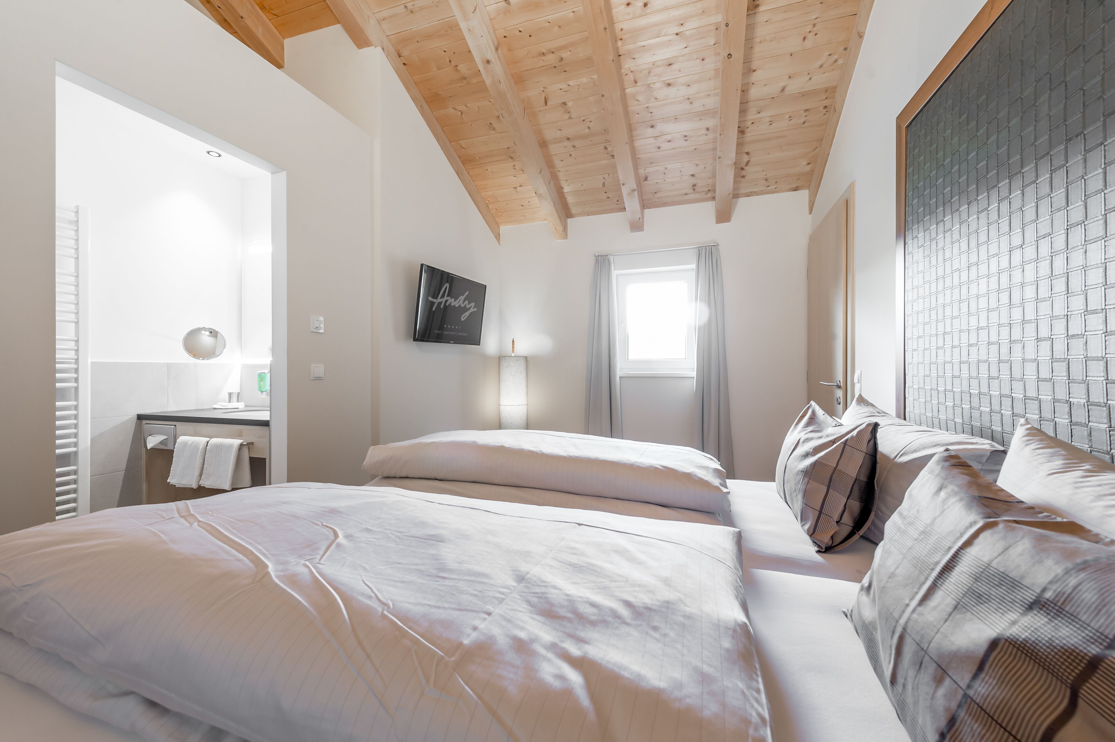 A bedroom with a wooden ceiling, a bed with white linens, a bedside table with a lamp, and a window with curtains.