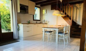 A modern kitchen with white cabinets, a dining area with a wooden table and chairs, and a staircase with wooden railings. - In Residence with swimming pool, Tennis, children's games 8 minutes from contis beach (Saint-Julien-en-Born)