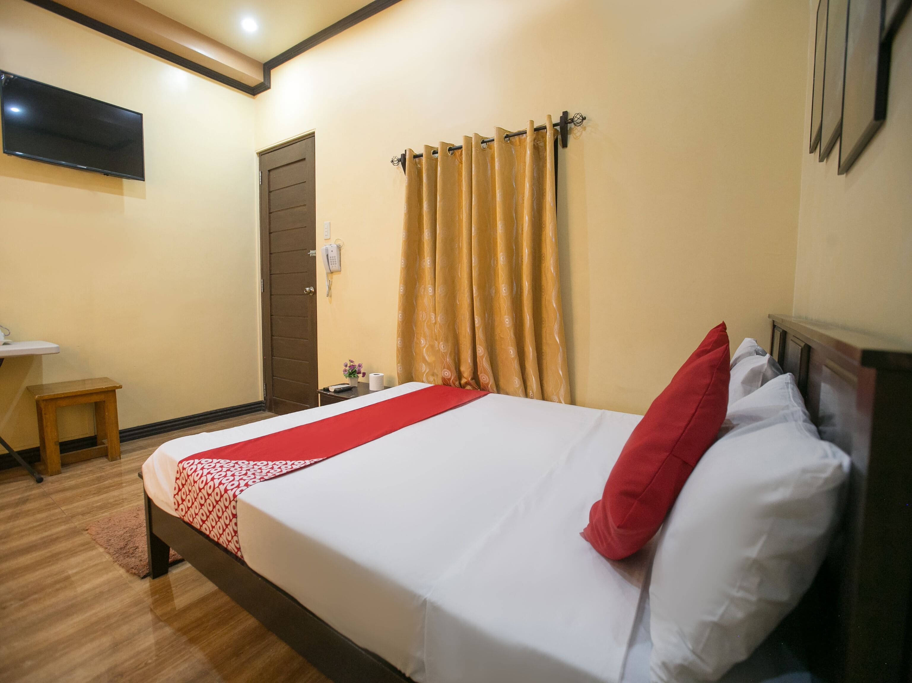 A neatly made bed with a red and white bedspread, a wooden nightstand with a telephone, and a wall-mounted TV.