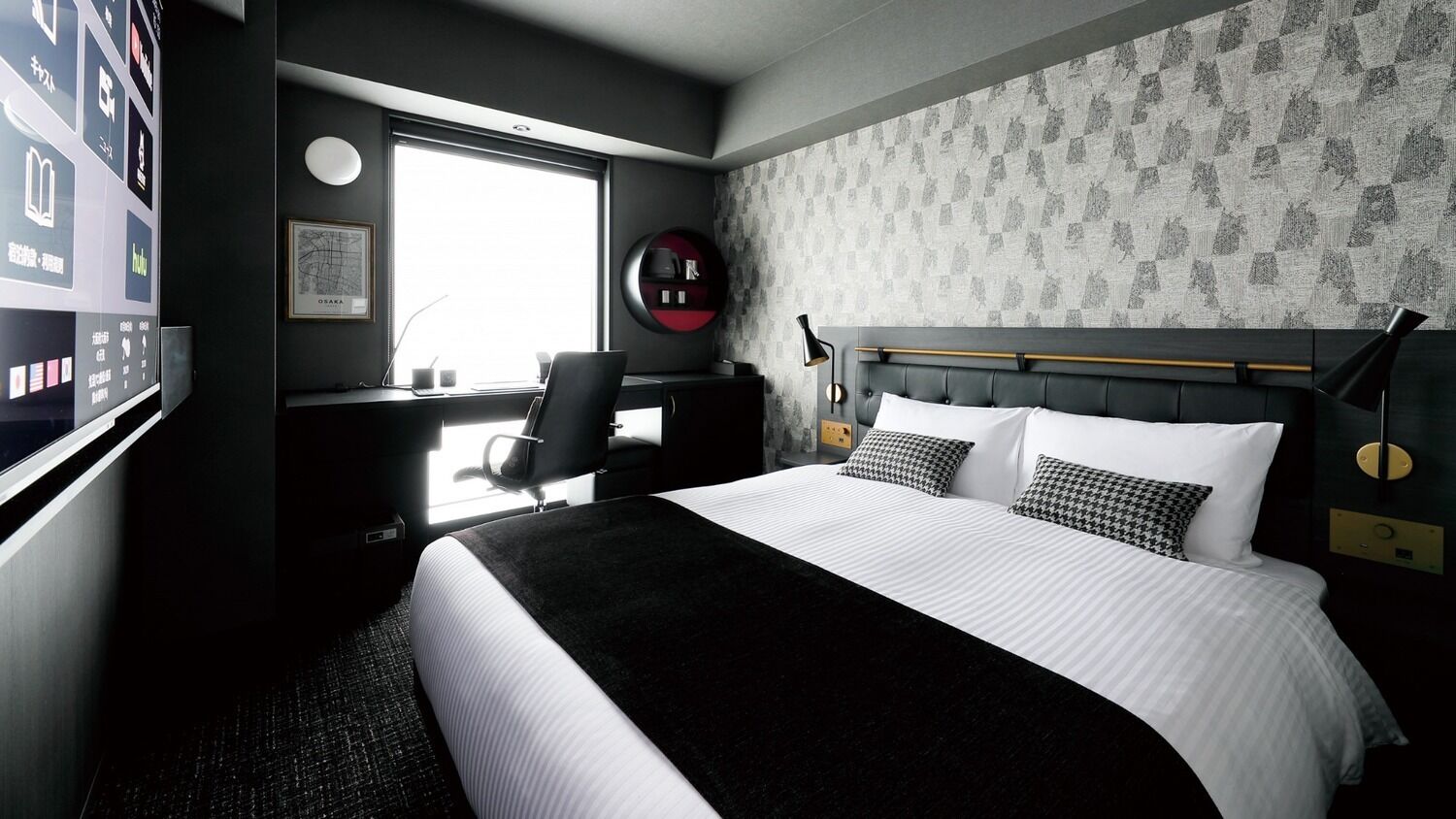 A hotel room with a bed, a desk, a chair, a window, and a wall with a patterned wallpaper.