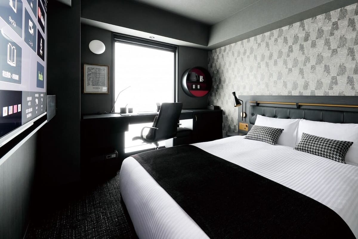 Ein Hotelzimmer mit Bett, Schreibtisch, Stuhl, Fenster und Wand mit ornamentalem Wandpapier.