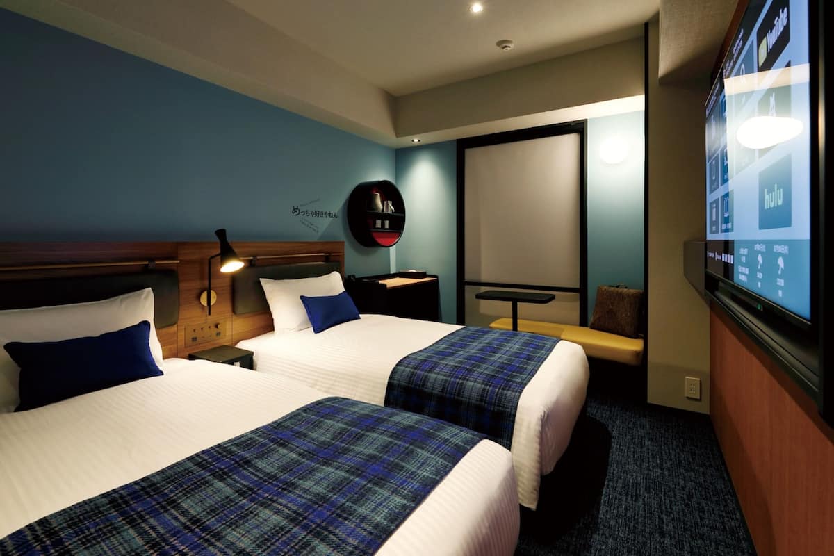 Ein Hotelzimmer mit zwei Betten, einem Fernseher, einem Schreibtisch und einem an der Wand befestigten Regal.