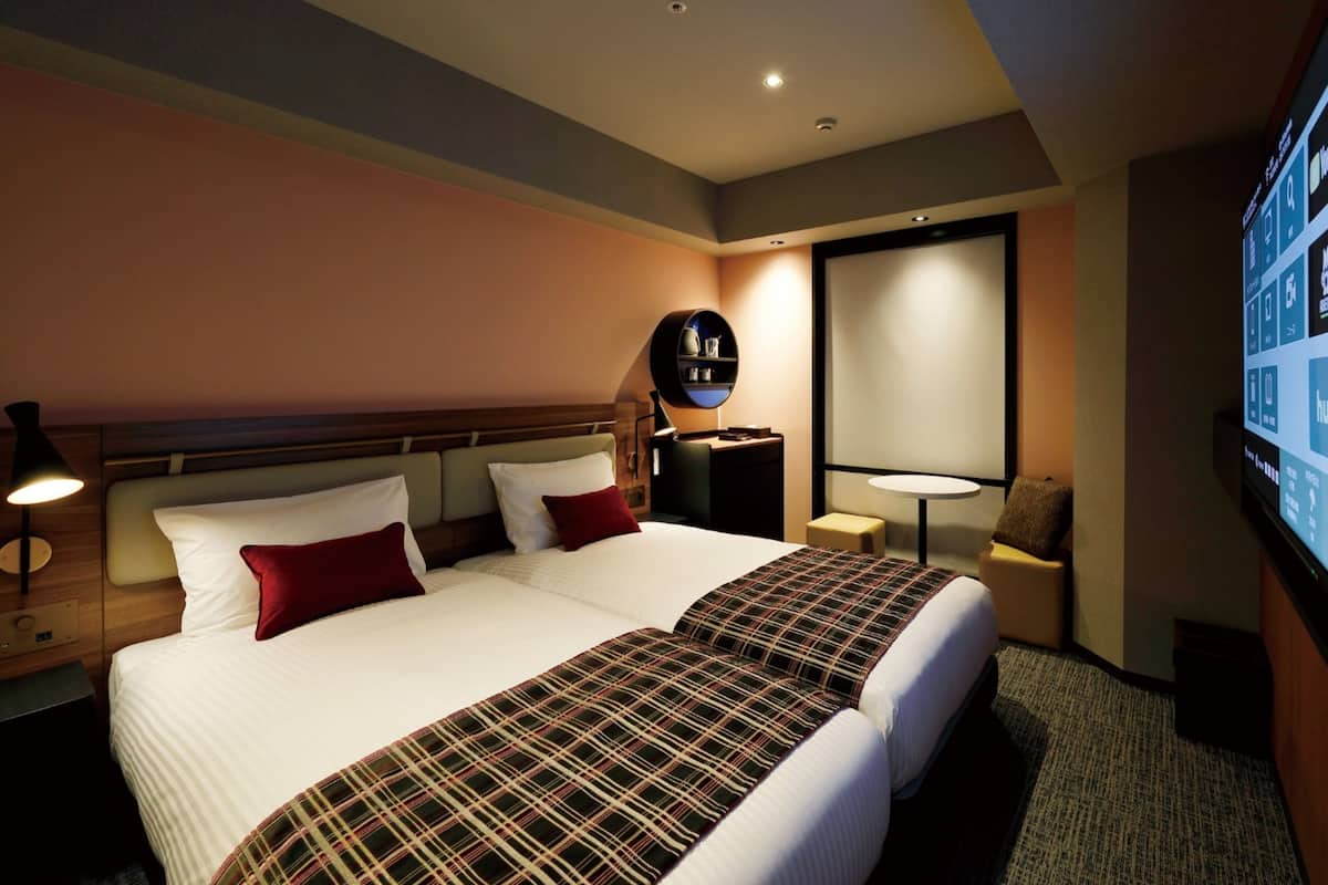 Ein Hotelzimmer mit zwei Betten, einem Nachttisch, einer Lampe, einem Fernseher und einem an der Wand befestigten Regal.
