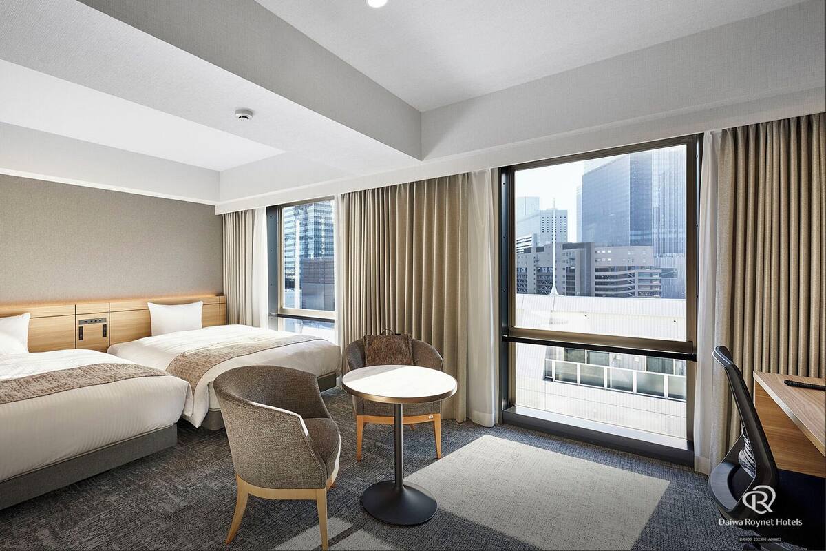 Ein Hotelzimmer mit zwei Betten, einem kleinen Tisch, einem Stuhl und Blick auf die Stadtlandschaft.