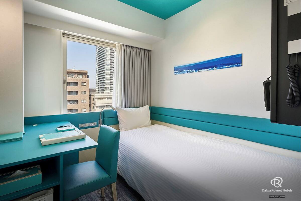 Ein Hotelzimmer mit Bett, Schreibtisch mit Stuhl, einem Fenster mit Vorhängen, einem Bild an der Wand und einem Telefon.