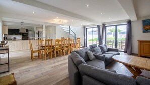 A spacious living area with a grey sectional sofa, wooden dining table, and chairs. A kitchen is visible in the background with a modern design.