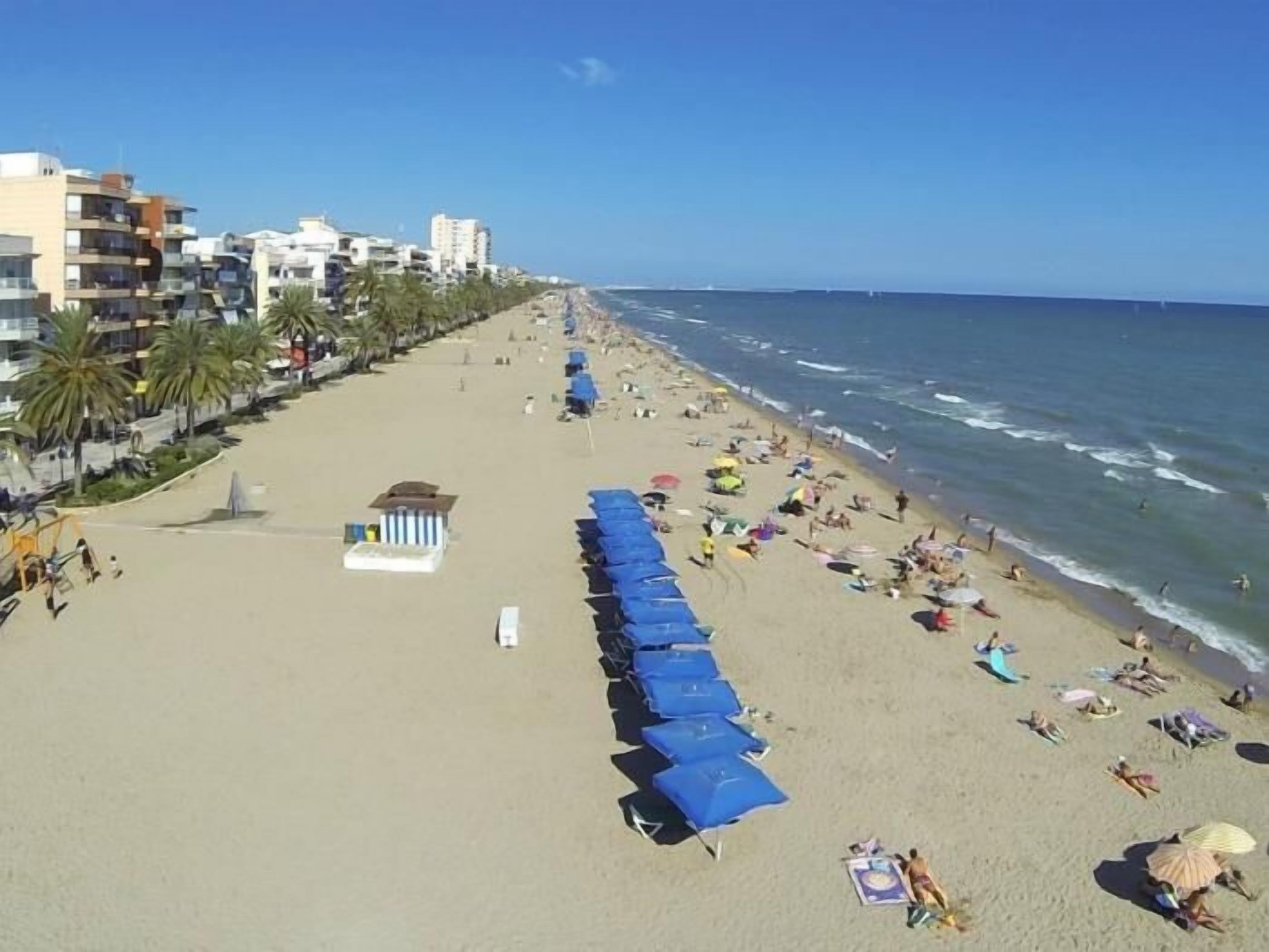 Une plage bordée de rangées de parasols bleus, une tour de surveillance pour les sauveteurs et des bâtiments en arrière-plan.