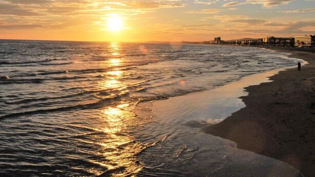 A beach with a sunset over the ocean, a person walking on the shore, and buildings in the background. - Apartament Suiza - Calafell (Calafell)