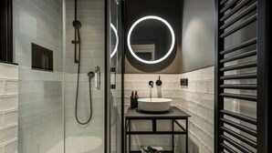 A modern bathroom with a glass-enclosed shower, a white sink with a black countertop, and a mirror with circular lighting. - THE ADAMS- Self Check in Hotel (Baden-Baden)