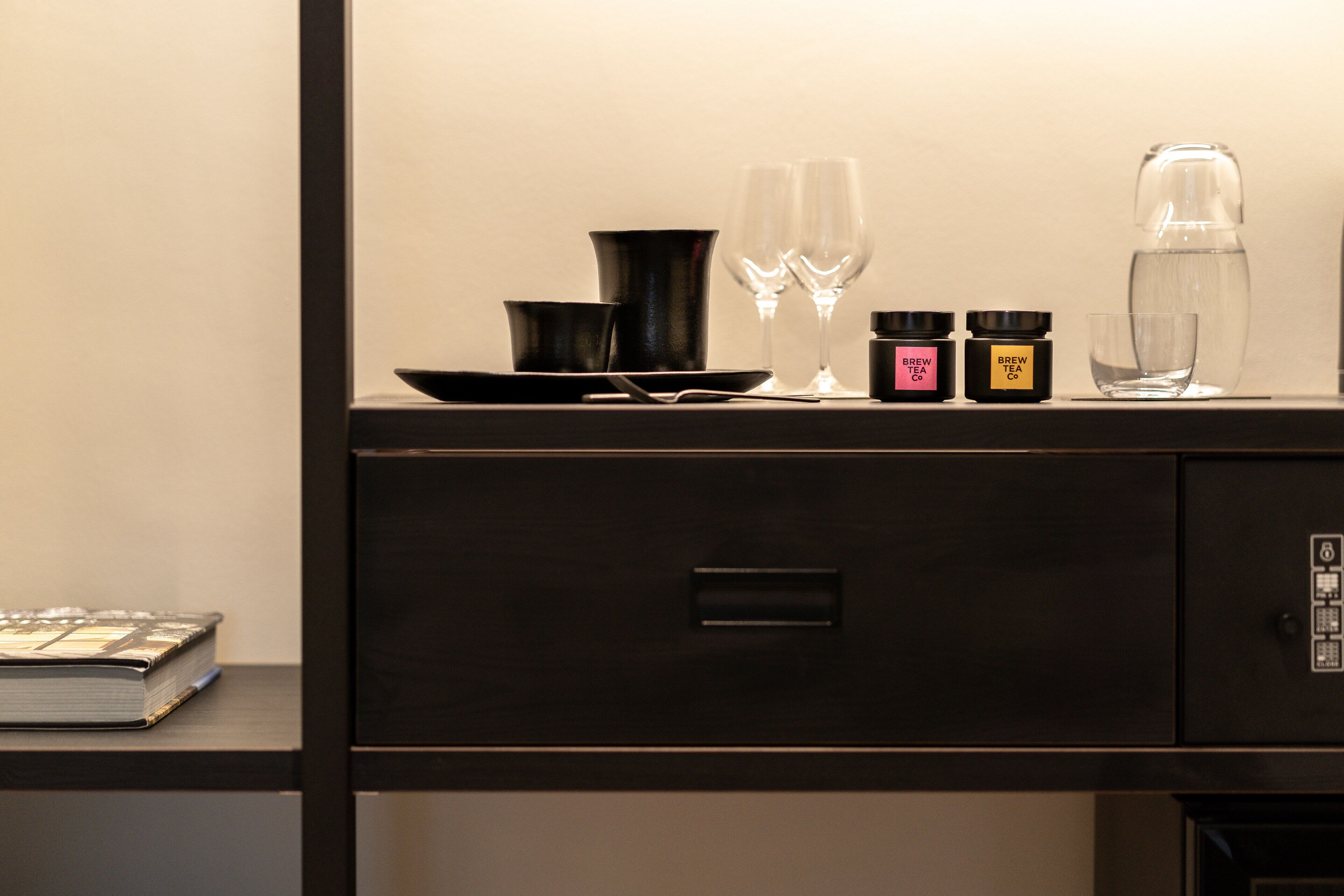 A neatly arranged desk with a book, a black tray, two wine glasses, a teapot, and two jars labeled "BREW TEA CO."