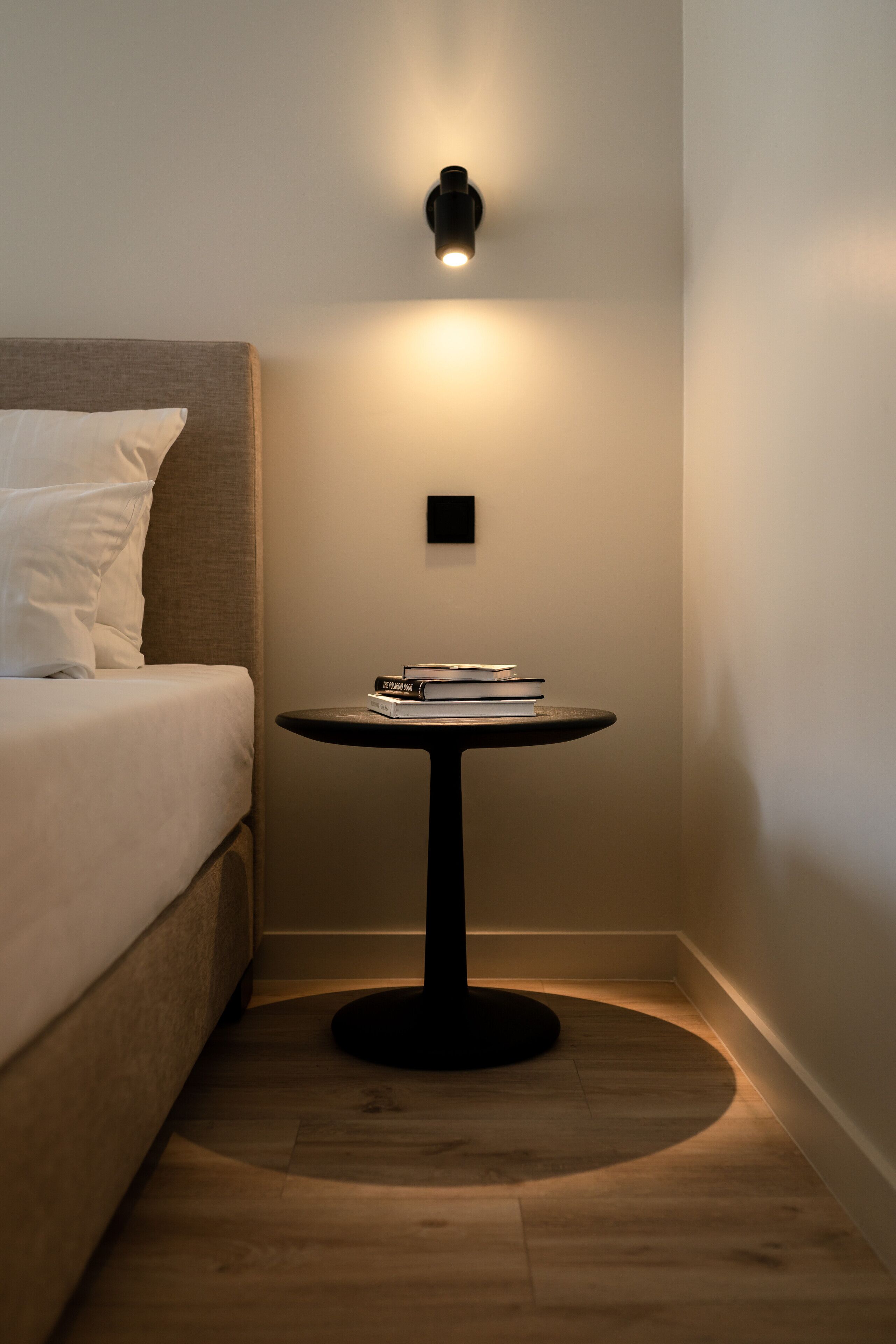 A hotel room with a bed, a small round table with books, a wall-mounted light, and a wall clock.
