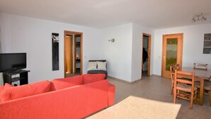 A living room with a red sofa, a TV, a dining table with chairs, and a kitchen area visible through an open door. - Apartment Lindamar - Calafell (Calafell)
