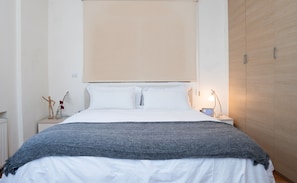 Una cama bien hecha con ropa de cama blanca y gris, una mesita de noche de madera con una lámpara y un armario de madera.