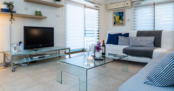A modern living room with a flat-screen TV, glass coffee table, sofa, and a mounted air conditioner.