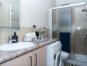 Un baño moderno con encimera de granito, un lavabo blanco, dispensador de jabón negro, inodoro blanco, cabina de ducha de cristal y una lavadora.