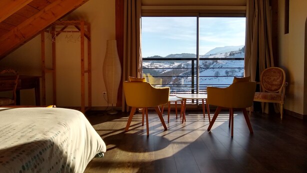 Een kamer met een bed, twee gele stoelen, een tafel en een uitzicht op een besneeuwd landschap door grote ramen.