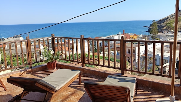 A terrace with two wooden lounge chairs and a large umbrella, overlooking a coastal town and the sea.