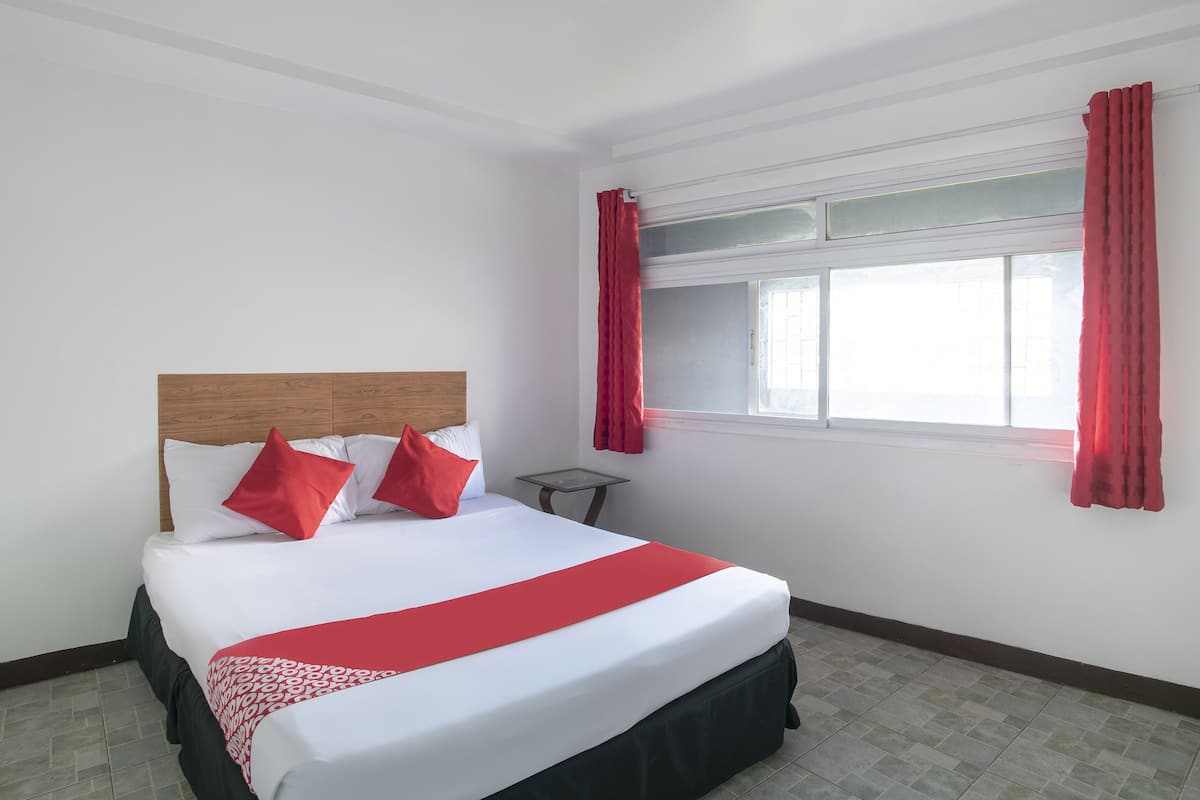 A hotel room with a wooden headboard bed, white bedding, red pillows, a red throw, and a window with red curtains.