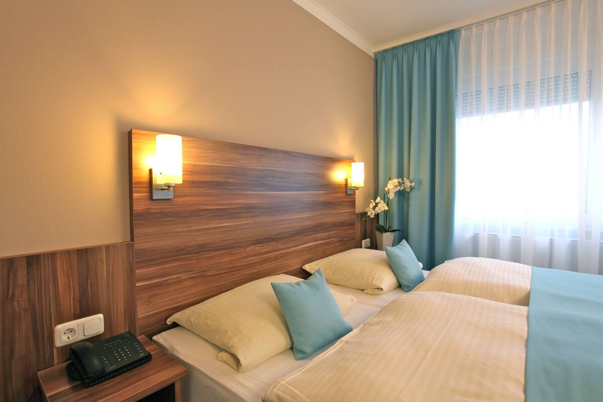 Une chambre d’hôtel avec deux lits, une tête de lit en bois, des lampes de chevet, une table de chevet avec un téléphone et une fenêtre avec des rideaux.