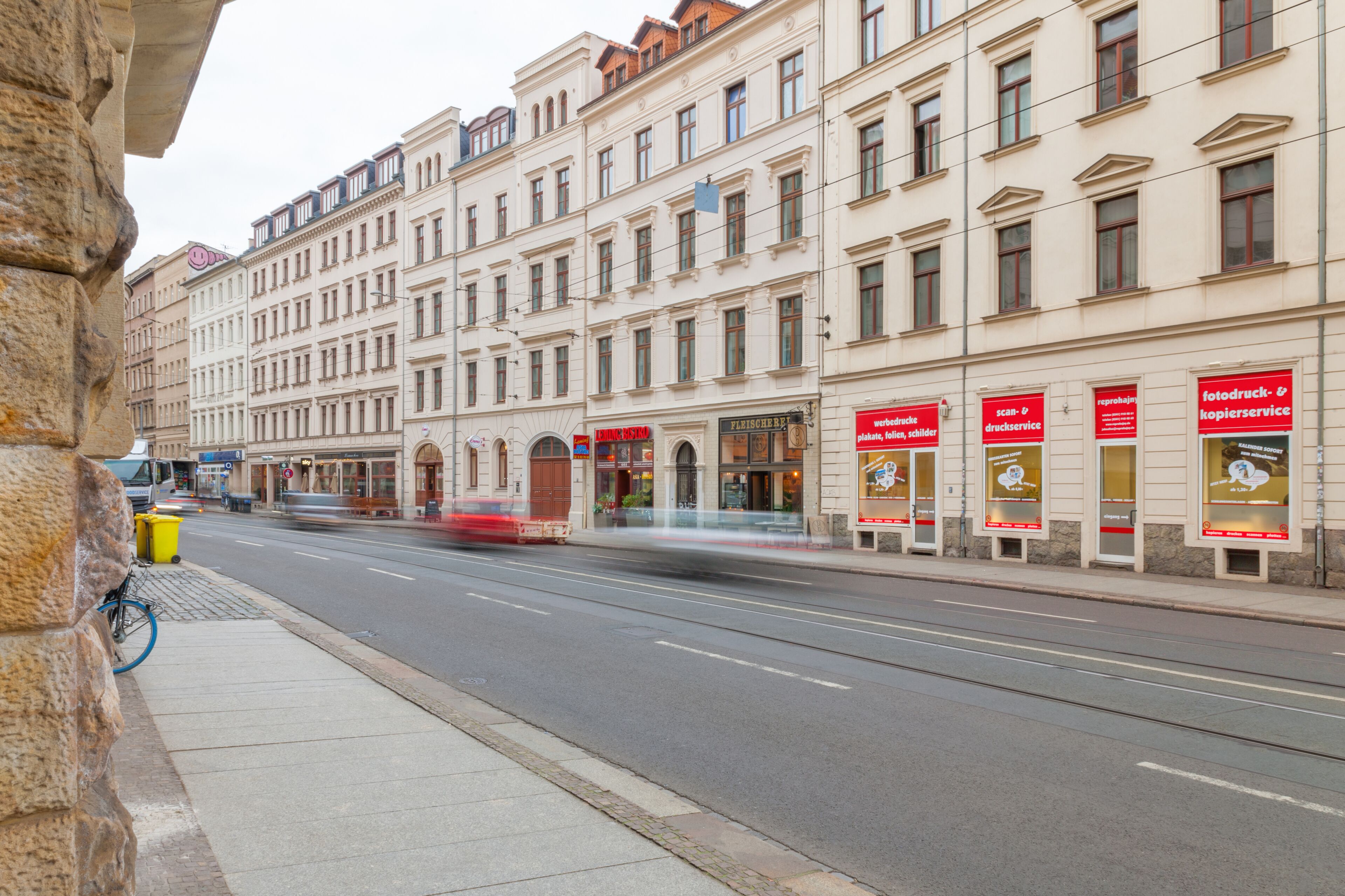 Eine Straße mit einer Straßenbahn, Gebäuden mit Beschilderung und einem Bürgersteig mit Mülleimer und Fahrrad.