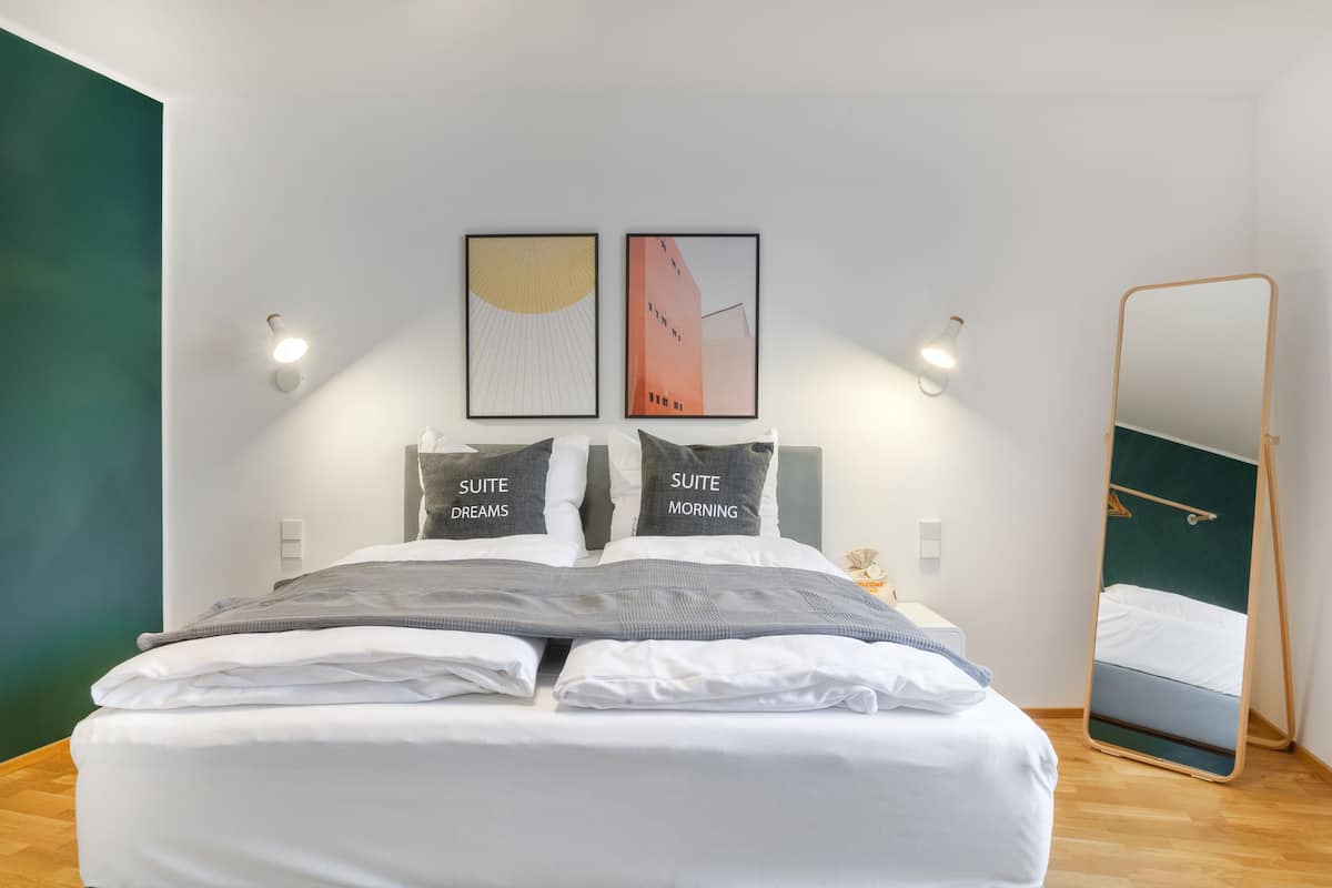 A modern hotel room with a neatly made bed, two framed artworks above the bed, a bedside table with a lamp, and a large mirror on the wall.