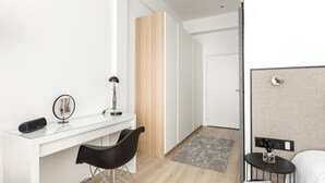 Une chambre d’hôtel moderne avec un bureau en verre, une chaise noire, un lit, une petite table avec un livre et un tapis gris.