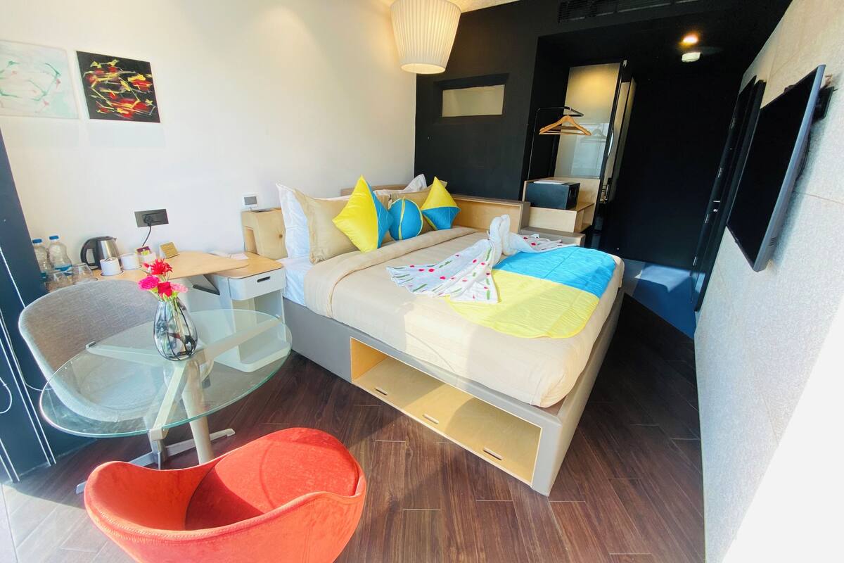 A modern hotel room with a bed, a glass table, a red chair, and a small desk with a lamp.