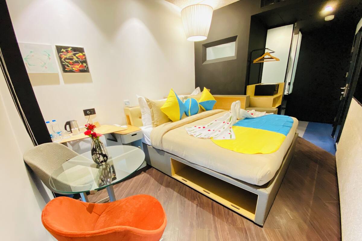 A modern hotel room with a bed, bedside table, chair, and a small desk with a glass top.