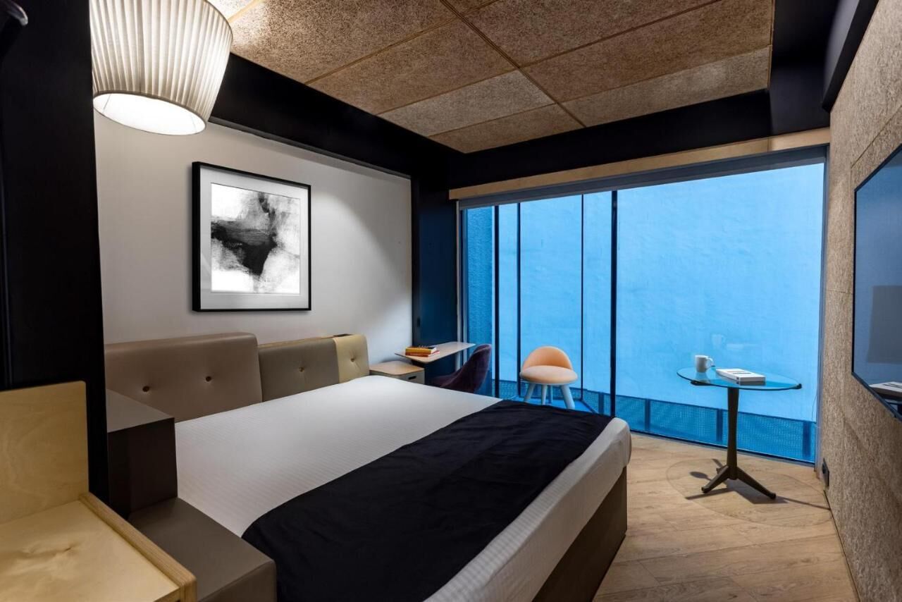 A modern hotel room with a large bed, a desk, a chair, a small table, and a large window offering a view.