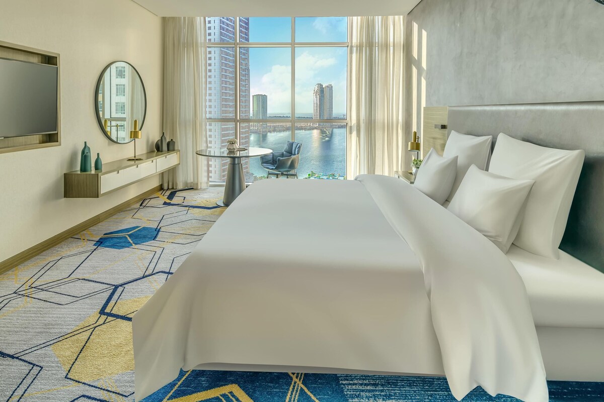 A hotel room with a large bed, a round mirror, a small table, a TV, and a view of a cityscape and water.