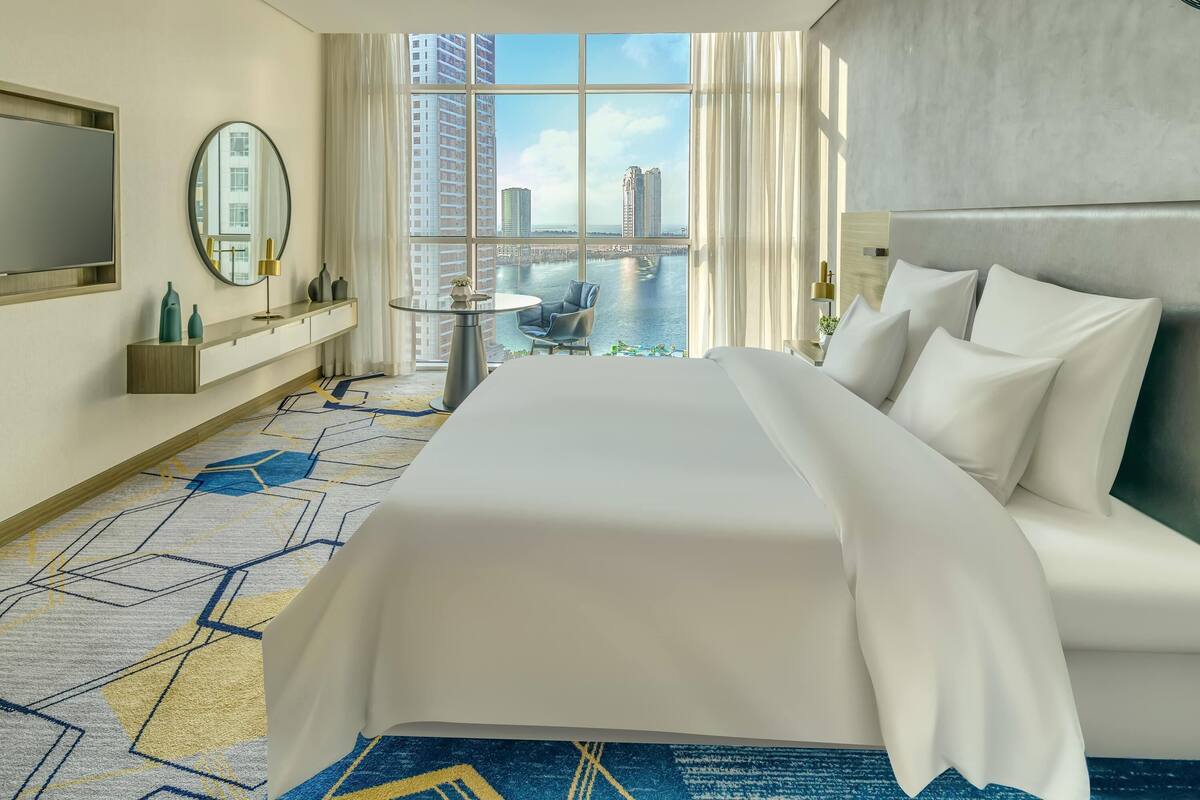 Habitación de hotel con una cama grande, un espejo redondo, una mesita, un televisor y vistas a la ciudad y al agua.