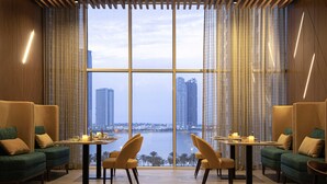 A restaurant with a view of a city skyline, featuring high-rise buildings, a body of water, and a modern interior with green sofas and wooden tables. - Pullman Sharjah (Sharjah)