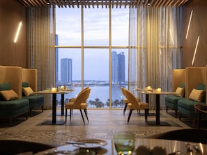 Ein Restaurant mit Blick auf eine Stadtlandschaft, einschließlich Wolkenkratzern, eines Gewässers und einem modernen Interieur mit grünen Sofas und Holztischen.