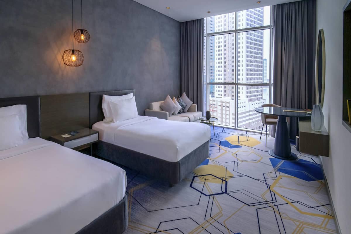 A modern hotel room with two beds, a large window with a city view, a desk, and a chair.