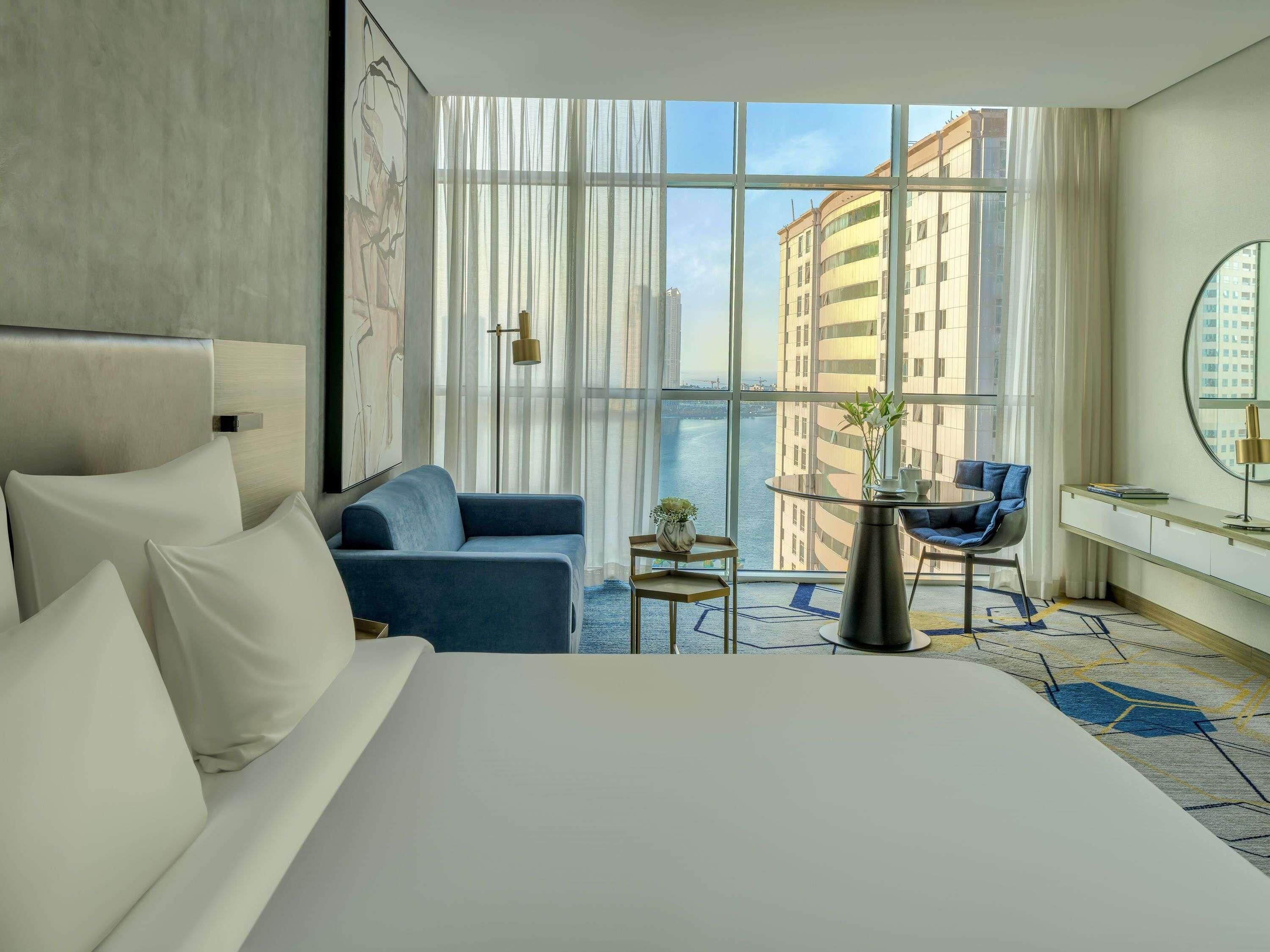 A hotel room with a large bed, a sofa, a small table, a chair, and a view of buildings through large windows.