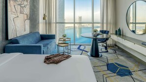 A modern hotel room with a large bed, a blue sofa, a small round table, and a view of the city and water. - Pullman Sharjah (Sharjah)