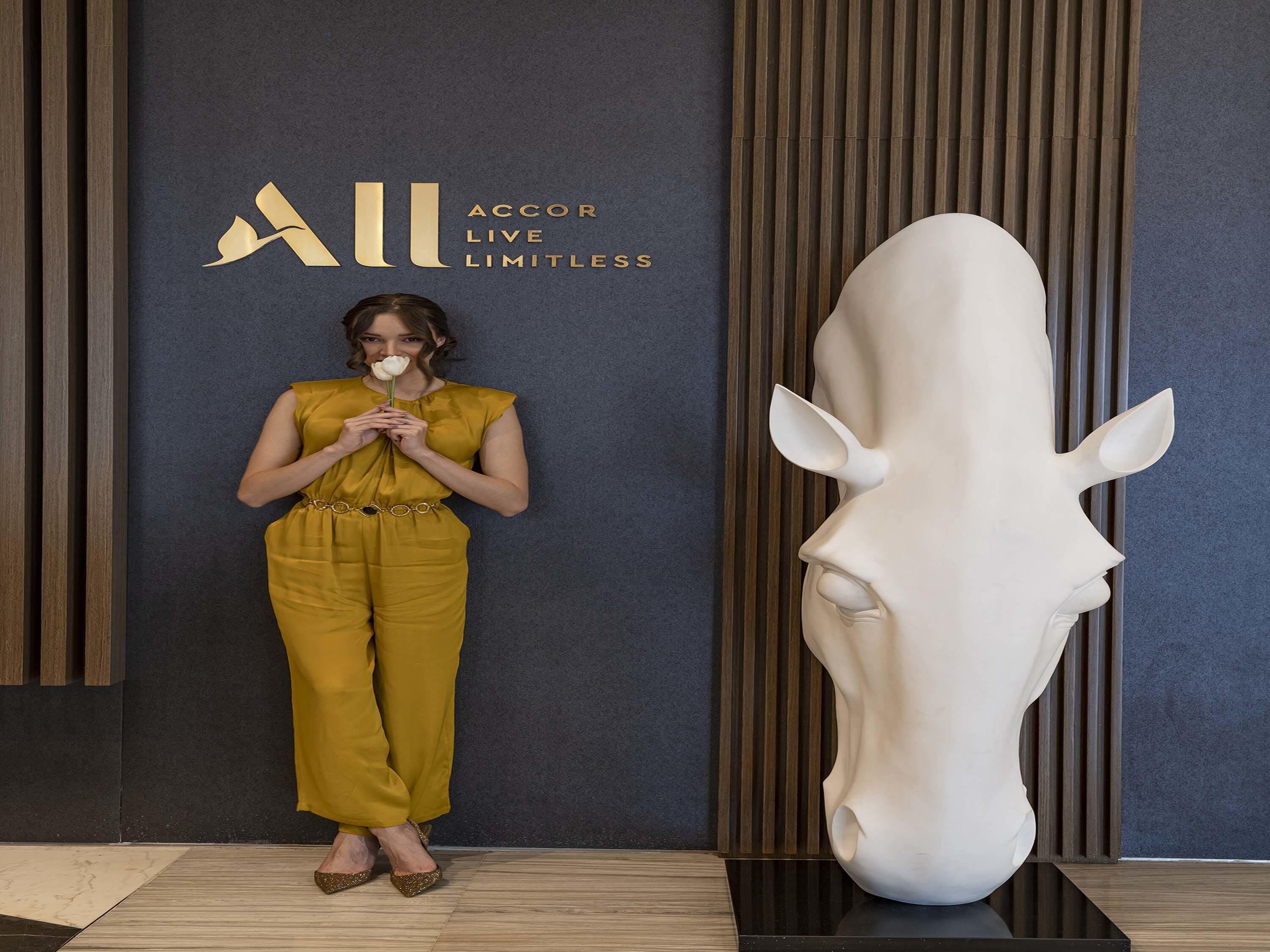 Una mujer con un mono color mostaza sostiene una rosa blanca, de pie junto a una gran escultura blanca de cabeza de caballo en un interior moderno con paredes oscuras y listones de madera verticales.