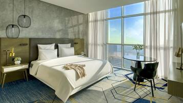 Quarto de hotel moderno com uma cama grande, uma mesa de cabeceira com abajur, uma pequena mesa com um vaso, e vista da cidade através de amplas janelas.